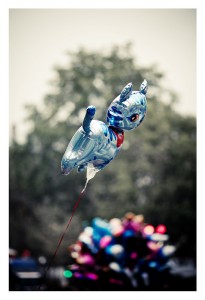 A shiny metallic blue balloon shaped like an animal floats in the air against a blurred background of greenery and colorful balloons, celebrating Techno Parade 2013. The balloon is tethered by a red string, marking the excitement of the 15ème édition.