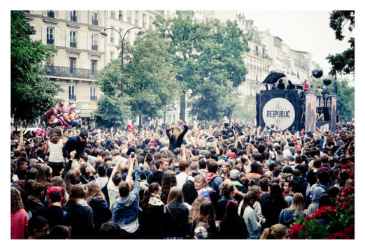 A large, energetic crowd gathers on a bustling urban street for the 2013 Techno Parade, with people dancing and cheering around a DJ stage labeled "REPUBLIC." The surrounding buildings provide a cityscape backdrop to the lively outdoor party scene at its festive 15ème édition.