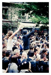 A large crowd of people, some standing on others' shoulders, enjoys the 15ème édition of Techno Parade in 2013. Many are holding up smartphones to capture the moment, with trees and buildings visible in the background.