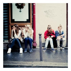 Four young people are sitting on a sidewalk against a building facade during the 15ème édition of the Techno Parade in 2013. Two young women on the left are looking to the side, another is looking at her phone, and a young man on the right is also looking to the side. There is graffiti on the wall behind them.
