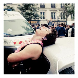 A person with short hair is leaning back against a car, eyes closed and mouth slightly open, wearing a pink floral lei. An ambulance is visible in the background, with a crowd of people gathered nearby. The scene appears to be outdoors during the 2013 Techno Parade, possibly La 15ème édition.