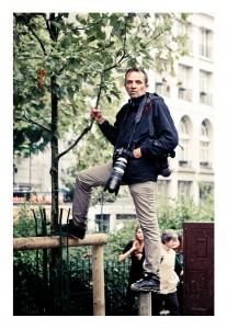 A person stands on a wooden post, holding onto a small tree for balance. They are dressed in a dark jacket and beige pants, with a large camera and telephoto lens strapped around their neck. In the background, people walk by a building with arched windows, reminiscent of the 2013 Techno Parade.