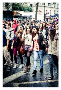 A group of people, primarily young adults, walk down a street during the 15ème édition of the Techno Parade in 2013. Some wear casual clothing, including jackets and jeans. The crowd appears engaged in conversation and the street is wet, possibly from recent rain. Buildings and balloons are in the background.