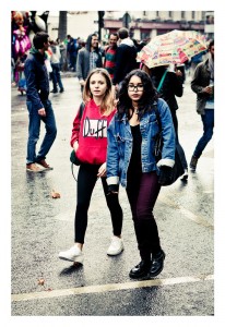 Two young women walking on a wet street during the busy 15ème édition of Techno Parade 2013. One wears a red hoodie with "Duff" printed on it and black leggings, while the other sports a denim jacket and dark pants. An umbrella decorated with colorful patterns is visible in the background.