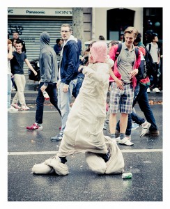 A person wearing an oversized padded costume that covers their entire body walks on a street during the 15ème édition of the 2013 Techno Parade. Humorously dressed, they catch the attention of onlookers, who watch with a mix of amusement and curiosity.