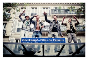 Four young men are standing on a glass-covered bus stop, dancing and pointing upward, likely inspired by the energy of the Techno Parade. They appear to be having a good time. A street sign reads "Oberkampf - Filles du Calvaire." In the background, there’s a building with ornate balconies and several windows.