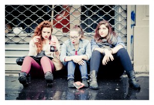 Three young women are sitting on the ground in front of a closed metal shutter at the 15ème édition of the Techno Parade. Each woman has long hair, wearing casual clothes and boots. They look relaxed, one is holding a drink, while the others are looking down or ahead. There's a plastic bag on the ground.