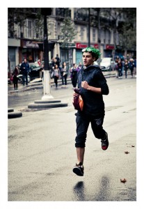 A person wearing a green leafy wreath, dark hoodie, and cropped pants runs through the city streets during the 15ème édition of the Techno Parade in 2013. A small crowd and several buildings are visible in the background on a cloudy day.