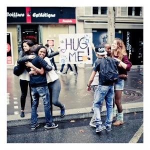 A group of people stands on a city street; two pairs are hugging, while another pair is preparing to hug. They hold a sign that reads "HUG ME!" with colorful handprints. The background shows storefronts and pedestrians, capturing the lively and cheerful essence of the 15ème édition of Techno Parade 2013.