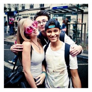 Three friends pose for the camera on a city street during Techno Parade 2013. The woman on the left sports large, round pink sunglasses and a gray tank top. The two men stand close on her right, one wearing glasses and a hoodie, the other a cap and printed T-shirt. All are smiling at the 15ème édition celebration.