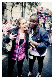 Two people smile and pose for the camera at the lively 15ème édition of Techno Parade 2013. One sticks out their tongue and flashes a peace sign while the other also shows a peace sign. The background is filled with other attendees, colorful balloons, and urban surroundings.