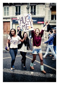 Three young people walk in the street; one holds a sign that reads "Hug Me!" The person on the right has their arms raised in excitement. They are surrounded by others participating in the 15ème édition of Techno Parade 2013 on a wet road with buildings and onlookers in the background.