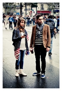 Two people stand on a rainy street during the 2013 Techno Parade. The woman on the left, holding a phone, wears a black leather jacket, a scarf with stars and stripes, jeans, and white boots. The man on the right, holding a drink, wears a brown jacket, striped shirt, scarf, and black jeans.