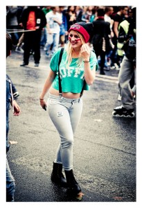 A young woman is walking outdoors during the busy 15ème édition of Techno Parade. She is smiling, wearing a green crop top with "OFF" printed on it, light-colored jeans, black boots, suspenders, and a black beanie with red and yellow writing. People are visible in the background.