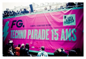 A large pink banner with "Techno Parade 15 Ans" printed on it is displayed prominently in front of a crowd, celebrating the 15ème édition. The banner also features logos and sponsors, including "FG." People are gathered around, watching from behind barriers and from a rooftop above.