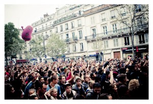 A large crowd of people gathered outdoors in a city street, many with hands raised, likely participating in the 2013 Techno Parade. In the background, multi-story buildings with balconies and trees line the street. A pink inflatable figure is seen hovering above the crowd during this 15ème édition.