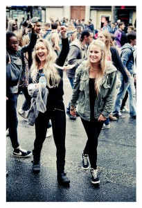 Two young women smile and walk among a crowd during the 15ème édition of the Techno Parade. The woman on the left raises her fist in the air while holding a water bottle in her other hand. People around them are engaged and moving, creating a lively atmosphere at this vibrant 2013 event.
