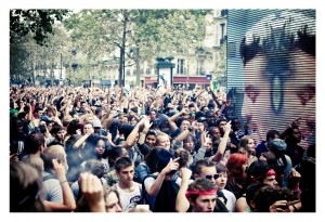 A large crowd of people gathered at the 15ème édition Techno Parade in 2013, many with their hands raised and some taking photos with their phones. A large screen in the background shows a close-up image of a face in monochrome. Trees and buildings are visible in the background.