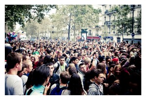A large crowd of people gathered outdoors during the daytime for the 15ème édition of the Techno Parade in 2013. Many are smiling, talking, and some have their hands raised. The background includes trees and buildings, creating a lively and festive atmosphere.