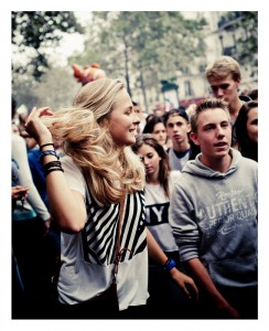 A crowd of young people are at an outdoor event during the 15ème édition of the Techno Parade in 2013. The focus is on a woman with long blonde hair, wearing a white shirt with a black abstract design, who is smiling and flipping her hair. The others are casually dressed, and trees and buildings are in the background.