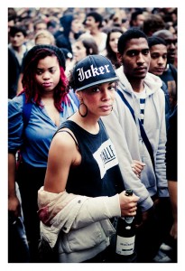 In the midst of the bustling 15ème édition Techno Parade, a central figure stands out in the crowd, sporting a black "Joker" cap and a black tank top with "MALLE" emblazoned on it. They hold a bottle in their left hand, a jacket draped over their arm, as others look around expectantly.