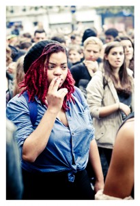 A person with red dreadlocks and wearing a black beanie and blue shirt smokes a cigarette in a crowded outdoor setting during La 15ème édition of the Techno Parade in 2013. The crowd consists of diverse individuals, some engaged in conversation and others looking around.