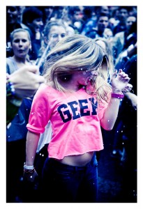 A young woman with long hair, wearing a bright pink crop top with the word "GEEK" printed on it, dances energetically in a crowd of people at La 15ème édition of the 2013 Techno Parade. Snow-like confetti is falling, and the atmosphere is lively and joyful.