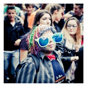 Amid the throngs at the Techno Parade 2013, a person in a crowded outdoor setting dons oversized blue sunglasses, a colorful wig, a red bow tie, and a plaid shirt. The vibrant attire makes them stand out among casually dressed revelers of this lively 15ème édition.