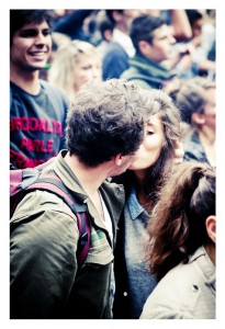 In the lively crowd of the 2013 Techno Parade's 15ème édition, a couple shares a romantic kiss. The man, with curly hair and a beard, sports a green jacket and red backpack, while the woman in a gray top embraces him. Around them, people are engaged and cheerful, adding to the festive atmosphere.