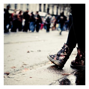 Close-up of a pair of legs wearing black floral boots standing on a wet pavement. In the background, a crowd of people is blurred, suggesting motion and activity at the 15ème édition Techno Parade in 2013. The weather appears overcast, and the ground is damp.