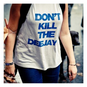 A person wearing a sleeveless shirt with the text "DON'T KILL THE DEEJAY" in bold blue letters stands out at the 15ème édition of the Techno Parade. The person is also sporting a wristband and bracelet, while another slightly blurred attendee can be seen in the background.