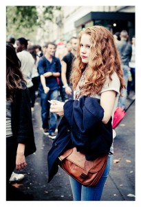A woman with long, curly red hair stands in a crowded street during the 15ème édition of the Techno Parade, holding a coat and a brown bag. She is wearing a light gray t-shirt and blue jeans, gazing ahead with a neutral expression. People and trees in the background suggest an outdoor setting.