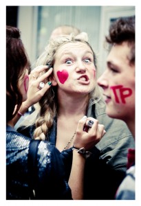 A young person with a braided hairstyle has a red heart and the letter "P" painted on their cheek, making a playful face at the 2013 Techno Parade. Another person beside them also has "P" painted on their cheek. A hand is touching the first person's face. The mood is lively and fun at this 15ème édition event.