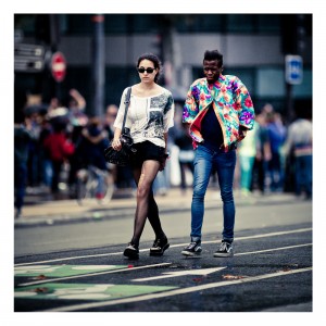 Two people walking on a city street during the vibrant 15ème édition of the Techno Parade 2013. The person on the left wears sunglasses, a white shirt, black shorts, and black stockings. The person on the right sports a brightly colored floral jacket and skinny jeans. A crowd and buildings are visible in the background.