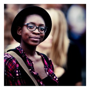 A person wearing glasses, a black hat, and a red checkered shirt smiles at the camera during the 2013 Techno Parade. The background is blurred, highlighting the person in the foreground.