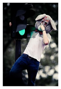 A person in a casual outfit and a large rabbit mask, possibly from the 2013 Techno Parade, stands with one hand holding onto a green traffic light, biting the ear of the mask. The background shows an out-of-focus outdoor setting with trees.