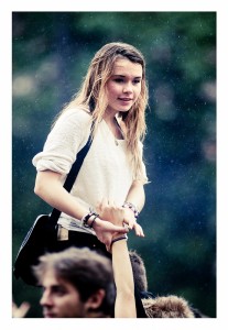 A young woman with wet hair and a white sweater is sitting on someone's shoulders outdoors in the rain at the Techno Parade 2013. She holds onto a raised hand while looking ahead thoughtfully. The background is blurred, showing greenery and raindrops.