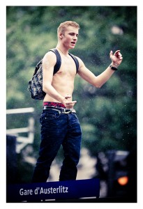 A young man with short hair stands shirtless, wearing blue jeans and a black backpack. He is making hand gestures and appears to be talking. The background is blurred with greenery. A sign below him reads "Gare d'Austerlitz," hinting at the vibrant energy of the 2013 Techno Parade, its 15ème édition.