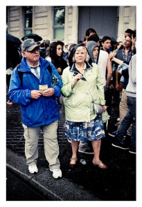 An elderly man in a blue jacket and cap stands next to an elderly woman in a light green raincoat and patterned dress. They are among a crowd of people, some of whom are wearing hoodies and jackets, standing on a wet pavement outside a building during the 15ème édition of Techno Parade 2013.