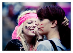 Two women are seen at an outdoor event during the 15ème édition of Techno Parade in 2013. One, with long blonde hair and a pink headband, has the word "FREE" written on her cheek. She is smiling and applying red face paint to the cheek of the other woman, who has brown hair. The background is blurry.