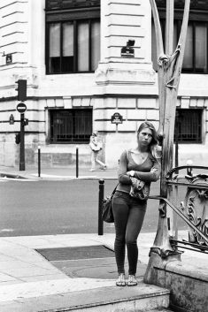 Le temps perdu ne se rattrappe plus A woman stands at the entrance to a subway station, leaning against a metal railing with her arms crossed, lost in temps perdu. A city street and building are visible in the background. Black and white photo.