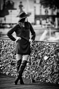 A woman in a chapeau, bottes de cuir, and robe noire stands on a bridge covered with love locks, gazing to the side. The black and white photo features an urban background softly blurred behind her.