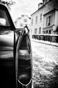 Close-up of the rear tail light of a vintage voiture de luxe parked on a cobblestone street, with blurred residential buildings in the background, in black and white.