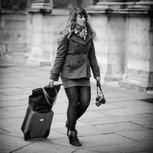 A woman walks along a stone-paved street, pulling a valise with one hand and holding a camera in the other. She wears a coat, scarf, patterned tights, and ankle boots. The photographe scene is captured in black and white.