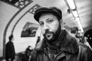 A bearded man wearing a cap, scarf, and leather jacket stands in a subway station, looking toward the camera. The background is blurred, showing other people and tiled walls. The image is in black and white.