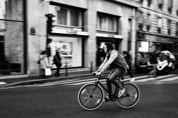 A toute vitesse A man wearing headphones rapidly rides a bicycle down a city street. The blurred background of pedestrians and storefronts emphasizes the vitesse and motion of the cyclist. The photo is in black and white.