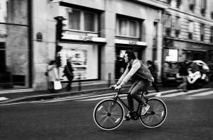 A man wearing headphones rapidly rides a bicycle down a city street. The blurred background of pedestrians and storefronts emphasizes the vitesse and motion of the cyclist. The photo is in black and white.