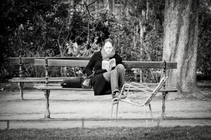 A woman sits alone on a park bench in Luxembourg reading a book, an empty chair in front of her. She is dressed warmly, with trees and bushes framing the scene. The black-and-white image evokes the essence of La lectrice.