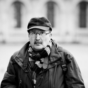 Black and white photo of Patrick Plazzi, an older man wearing glasses, a cap, a striped scarf, and a winter jacket, looking slightly past the camera with a neutral expression. The background is blurred.