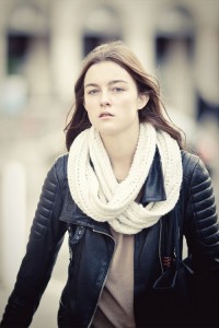 A young woman with long brown hair showcases her natural beauty in a black leather jacket over a beige top and thick white knit scarf. She stands outdoors, looking forward with a neutral expression against a softly blurred background.